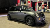 A dark gray MINI Cooper with black wheels is parked at a Synergy-branded gas station at night. The car features a hood scoop and sporty design elements. The background includes gas pumps and a dimly lit forecourt. The license plate reads "WA55XXX" (partially obscured for privacy).