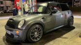 A gray MINI Cooper with black racing stripes and aftermarket wheels is parked at a gas station at night. The car is positioned next to a red and white Synergy fuel pump. The surrounding area is dimly lit, highlighting the MINI and part of the gas station structure.