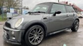 A silver MINI Cooper is parked on a wet pavement near a blue fence and an auto service sign. The car has black accents, a sunroof, and aftermarket alloy wheels. Overcast sky in the background. Another car and industrial structures are visible in the distance.