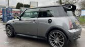 A metallic gray MINI Cooper is parked on a wet pavement with aftermarket large alloy wheels and a prominent rear spoiler. The car is positioned at an angle, showcasing its side and rear. A tree and a building with signage are visible in the background on a cloudy day.