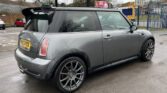 A grey MINI Cooper hatchback is parked on a wet road in an industrial area. The car features a black roof, large silver alloy wheels, and a rear spoiler. The license plate reads "NG53 PYX". Buildings and a fenced area with a gate are visible in the background. The sky is overcast.