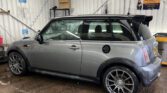A metallic gray MINI Cooper with racing wheels is parked inside a garage with a corrugated metal wall and a wooden floor. Cleaning equipment is visible on the left, and a sign with orange lettering is mounted on the wall. The MINI's surface is slightly wet, indicating recent washing.