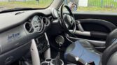 The interior of a right-hand drive MINI Cooper is shown, featuring a black and blue color scheme. The dashboard includes various dials and controls. The steering wheel is on the right with an air freshener hanging. Leather seats with blue fabric inserts and a manual gear shift are visible.