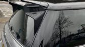 A gray MINI Cooper S hatchback is parked in a lot. The rear window reflects a tree and nearby structures. The car features a roof spoiler and a partially visible UK license plate. In the background, other parked cars, a billboard, and a fenced area with a building are visible.