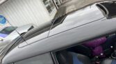 A black MINI with a sunroof is parked outside a building with a sign on the front. A person wearing a purple jacket is seated in the passenger seat, partially obscured. Other vehicles are visible in the background near the building, and the area appears to be a parking lot.