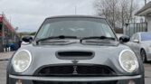 A metallic dark gray MINI Cooper with license plate VN53 PXX is parked on a wet pavement. The car has prominent headlights, a black bonnet scoop, and the MINI emblem on the grille. The background features a gray sky, leafless trees, and additional parked vehicles, including a silver car to the right.