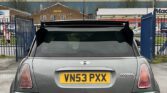A rear view of a gray MINI Cooper with the registration plate VN53 PXX is situated in a parking area under an overcast sky. Blue gates flank the car, leading to a building in the background with signage reading "Hand Car Wash" and "Mac." The surroundings hint at an industrial area.
