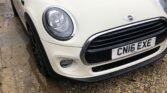 A white MINI Cooper is parked on a driveway made of pebbles and stone tiles. The license plate reads "CN16 EXE." The car features distinctive round headlights, a black grille, and black side mirrors. The surrounding area includes bushes and a wooden fence in the background.