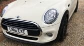 A white MINI Cooper with a black roof is parked on a gravel driveway beside a brick wall covered with greenery. The car's license plate reads "CN16 EXE." In the background, there is a wooden fence and a pink house. The headlights and front grille are prominently visible.