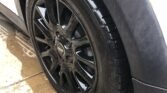 Close-up of a black vehicle tire on a white MINI. The tire is clean and shiny, indicating it's newly washed. The wheel rim is glossy black with a five-spoke design. The car is parked on wet pavement, with water droplets visible on the ground beside the tire.