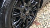 Close-up image of a shiny black car wheel and tire, showcasing the detailed and reflective surface of the alloy rim. The word "MINI" is prominently displayed in the center cap. The tire tread and sidewall markings are visible, with small pebbles and stones scattered on the ground beneath.