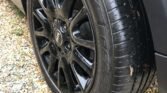 Close-up of a car tire and black alloy wheel on a gravel driveway. The tire appears to be new, with clean, shiny tread and visible tire branding. Part of the vehicle's white body and black wheel arch can be seen, typical of a stylish MINI. A few small leaves and pebbles are scattered around the tire.