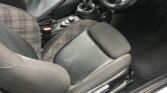         Interior of a MINI compact car featuring two black and gray plaid fabric front seats with black leather bolsters. The dashboard includes a large circular central display and multiple dials. The steering wheel has a silver logo in the center. A manual gear shift is visible between the seats.