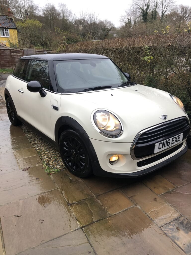 A white MINI Cooper with a black roof and black rims is parked on a wet, paved driveway next to a grassy garden. The car's headlights are on, and there is a license plate reading "CN16 EXE" at the front. Trees and a yellow house with a red roof are visible in the background.