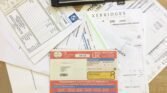 A tabletop spread with various papers including vehicle documents, invoices, and an owner's handbook partially obscuring other documents. A set of car keys rests on a black key wallet at the top. A UK vehicle registration certificate is prominent. Feet are visible at the bottom edge, beside a MINI brochure.