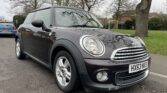 A black MINI Cooper parked on the side of the road with leafless trees and a grassy area in the background. The car has a white license plate with the registration "HX63 NXS." The sky is overcast, and another blue car is parked further down the road.