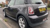 A black MINI Cooper is parked on the side of a residential street. The car has silver alloy wheels and a license plate reading "HX63 MXS." Houses and trees are visible in the background. The rear and side view of the MINI highlights its compact design and distinctive taillights.