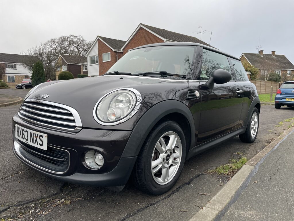A black MINI Cooper with silver detailing is parked on the side of a suburban street. The car has a UK number plate reading "HX63 NXS." Houses and trees are visible in the background, with a cloudy sky overhead. The vehicle is viewed from the front left side.