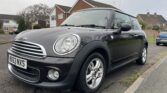 A black MINI Cooper with silver detailing is parked on the side of a suburban street. The car has a UK number plate reading "HX63 NXS." Houses and trees are visible in the background, with a cloudy sky overhead. The vehicle is viewed from the front left side.