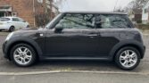 A black MINI Cooper is parked on the street in a residential area. The car features silver alloy wheels and a dark-tinted rear window. Brick houses and bare trees are visible in the background, with a white car parked nearby. The weather appears overcast, and the street is dry.