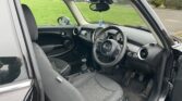 Image shows the interior of a MINI compact car with black seats and upholstery. The driver's seat and steering wheel are prominent, and the dashboard features various controls and a small screen. A hanging air freshener is present. Through the window, green grass and residential houses are visible.
