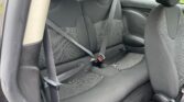 The image shows the backseat of a compact MINI car with black upholstery. The seat has a gray and black patterned fabric in the middle and includes three headrests. Two seat belts, one fastened and one unfastened, are visible. The image also shows a bit of the front passenger seat and a green lawn outside.