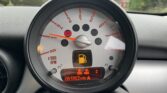 Close-up of a MINI car dashboard displaying a round tachometer with redlines starting at approximately 6,000 RPM. A fuel gauge symbol and 61,862 miles appear on the digital odometer below. Multiple dashboard warning lights are illuminated, indicating potential issues. The background shows a blurred view of parked cars and greenery.