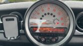 A car dashboard featuring a central speedometer with readings up to 160 mph, characteristic of MINI design. Below it, a digital display shows radio information. Control buttons for audio and car functions are directly underneath. To the left of the speedometer is a mounted phone holder labeled "Acleane." Houses are visible outside.