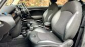 The interior of a MINI car showcases the front seats and dashboard. Black and gray leather seats with headrests. Manual gear stick, cup holders, and center console are visible. The dashboard features standard controls and a steering wheel on the left side. Outside, brown foliage is seen.