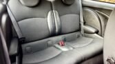 The image shows the backseat of a MINI compact car. The seats are upholstered in a mix of black fabric and leather. Two headrests are visible, along with seatbelt buckles and a fold-down armrest at the center. The car doors and interior side panels feature a sleek, modern design in the same color scheme.