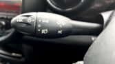 Close-up of a MINI's steering column showing the turn signal lever with various symbols. The lever includes controls for turn signals, high beams, and possibly other functions. The dashboard and part of the vent are partially visible in the background.