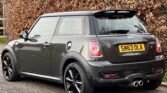 A dark grey MINI Cooper parked on a road with autumn leaves on the bushes in the background. The car has black alloy wheels, a rear spoiler, dual exhaust pipes, and the registration plate "SM63 OLA." The windows are slightly tinted, and the car is captured from a rear side angle.