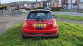 A red MINI Cooper with a black roof and black racing stripes is parked on a grassy area. The license plate reads "MT11 ZVA." In the background, there is a row of brick houses, a street, and a fenced property. The sky is clear, suggesting it's a residential neighborhood.