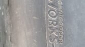 Close-up image of a black MINI car part with the words "JOHN COOPER WORKS" embossed in the center. The text is enclosed in an oval shape, with the words "JOHN COOPER" positioned vertically to the right of "WORKS." The surface around the text appears slightly worn.