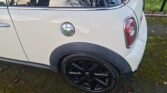 Close-up of the rear side of a white MINI Cooper parked on a mossy surface. The image shows the back wheel, part of the rear bumper, and the gas cap. The black roof and tinted window reflect bare tree branches against the sky, while fallen leaves surround the vehicle.