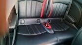 The image shows the back seat of a black leather MINI interior with red accents. The seat design includes vertical stitching and a prominent red strip in the center. There are seatbelt buckles and a central armrest/cupholder folded down. The car door on the right is partially visible.