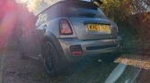 A silver MINI Cooper S with black accents is parked on a gravel path. The car, an R56 model featuring a JCW kit, boasts a spoiler and black alloy wheels. Sunlight filters through clouds in the background, creating a lens flare effect over the vehicle. Lush green foliage surrounds the area. The license plate reads "KY07 SCR.