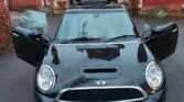 A black MINI Cooper with its front doors and rear hatch open is parked on a driveway in front of a historic-looking brick and stone building. The vehicle's license plate reads "GK13 FDV." The building features archways, decorated windows, and a red brick exterior.