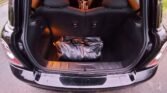 View of a MINI's open trunk showing the back seats and the interior. The trunk contains a single black duffel bag positioned in the center. The car is parked on an asphalt surface, and the interior lights are on, illuminating the trunk space.