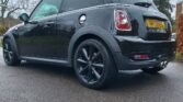 A black MINI Cooper SD with tinted windows is parked on a paved surface. The car has a two-door hatchback design, black alloy wheels, and a rear spoiler. The background features bare-branched trees, a hedge, and a cloudy sky. The vehicle's license plate is visible but partially obscured.