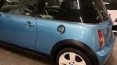 A blue MINI Cooper S with a white roof and wheels is parked in an indoor garage. The car's tinted windows and an "R53" decal on the rear quarter window add to its character. The well-lit garage also reveals another white vehicle partially visible in the background.