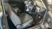 The interior of a compact MINI car featuring cloth-covered front seats, a dashboard with various controls, a steering wheel, and a speedometer with a white face and orange needle. The driver seat shows some wear, and the floor appears a bit dirty, especially near the pedals.