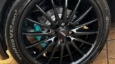 Close-up image of a car's black alloy wheel with a multi-spoke design and a visible blue brake caliper behind it. The tire, branded "Ventus V12 evo2," features prominent tread patterns and is mounted on a wet, textured brick surface. This stylish wheel is perfectly suited for your MINI.