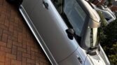 A silver MINI Cooper with a black roof is parked in a residential driveway. The car has black wheels and side mirrors, and it is positioned at a slight angle, showing the driver's side. The brick-paved driveway complements the sleek MINI, with bushes and charming brick houses in the background.