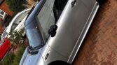 A silver MINI Cooper with a black roof and black wheels is parked on a brick driveway in front of a suburban house with greenery and a red car nearby. The vehicle’s sleek design and modern appearance are highlighted from a side view in the image.