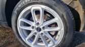 Close-up of a vehicle's right front wheel featuring a Michelin Primacy 4 tire and a multi-spoke MINI wheel rim. The tire and rim are clean and in good condition. The background shows part of the car's dark-colored fender and a gravel ground surface.