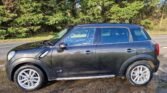 A black MINI Cooper Countryman is parked on a gravel surface beside a road. The car's exterior is shiny, with silver alloy wheels glinting in the sunlight. The background includes a row of green trees and bushes under a clear sky. Viewed from the side, the vehicle showcases its sleek four-door design.