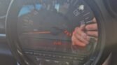 Close-up of a MINI car dashboard showing a speedometer with a maximum of 140 mph and a digital clock displaying 21:32. Hands are captured in the reflection on the plastic gauge cover. The speedometer needle is at 0 mph, and the surrounding landscape with trees and a cloudy sky is faintly visible through the window.