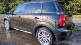 A dark gray MINI compact SUV is parked on a wet, gravel surface next to a road. The car has four doors, silver alloy wheels, and tinted windows. The rear lights are red, and the vehicle's reflection can be seen in a puddle on the ground. Tall green trees line the background.