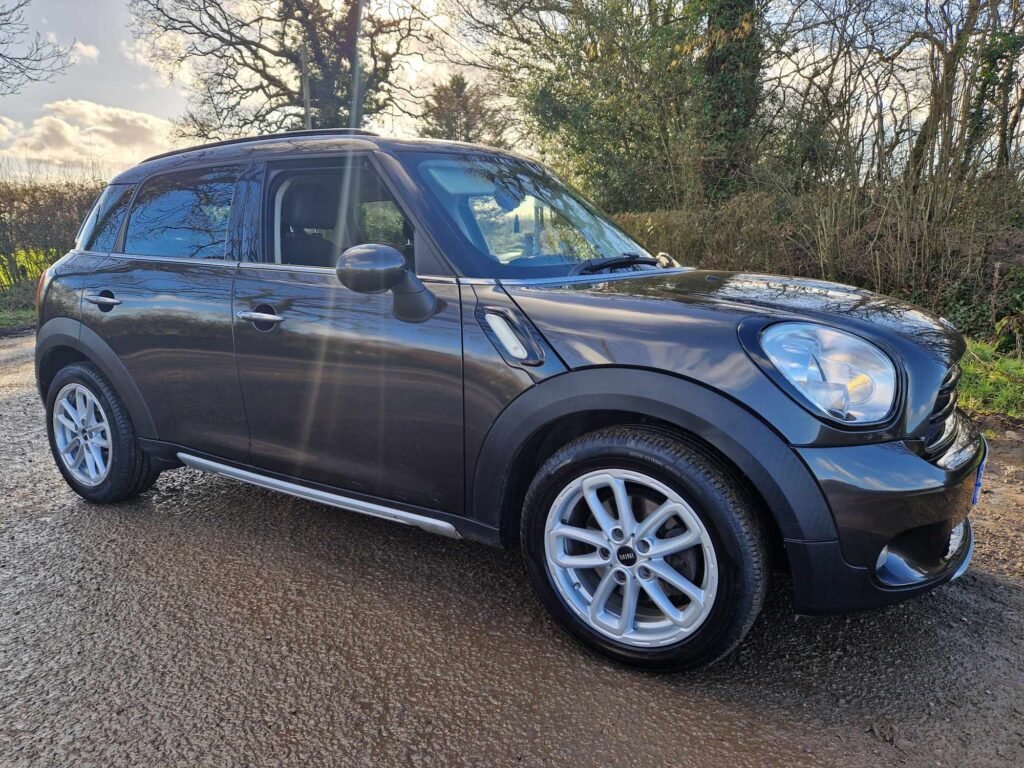 A dark-colored MINI compact SUV is parked on a muddy, rural path surrounded by leafless trees and bushes. The vehicle’s body is glossy, reflecting the bright sunlight filtering through the cloudy sky. The SUV features five doors, silver alloy wheels, and chrome side mirrors. The ground is wet, suggesting recent rain.